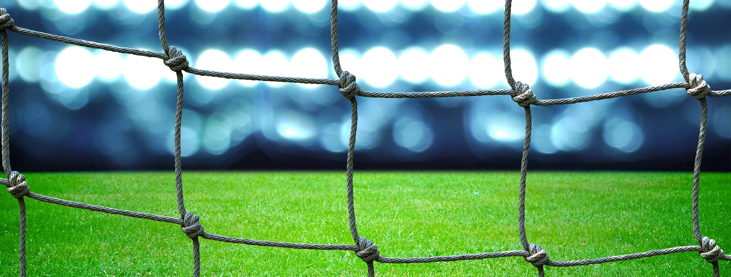 Rede de gol em primeiro plano, com gramado verde e refletores acesos desfocados ao fundo