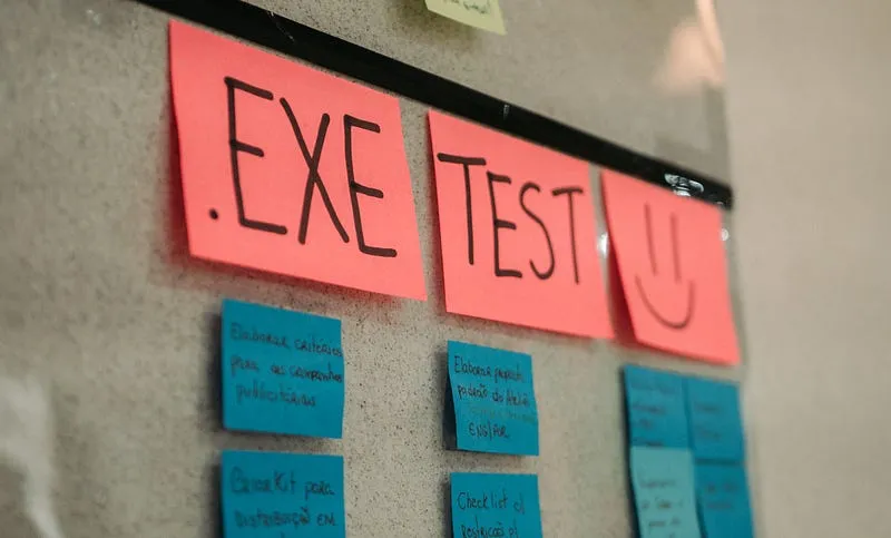 Quadro de cortiça com post-its: na linha de cima, três post-its vermelhos com ".EXE", "TEST" e um rosto sorridente; abaixo, post-its azuis com tarefas escritas à mão