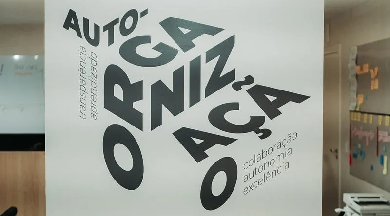 Painel em uma parede de escritório com a palavra “auto-organização” em letras grandes e, ao redor, as palavras transparência, aprendizado, colaboração, autonomia e excelência