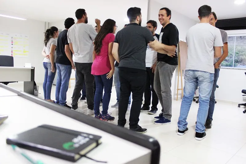 Grupo de pessoas de pé, conversando em roda num escritório, com um caderno sobre a mesa em primeiro plano