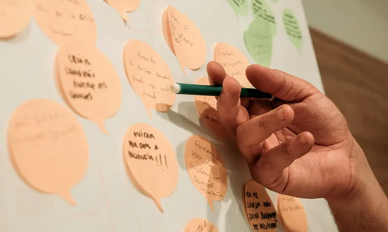 Mão segurando uma caneta verde escreve em post-its em forma de balão de fala colados em uma parede