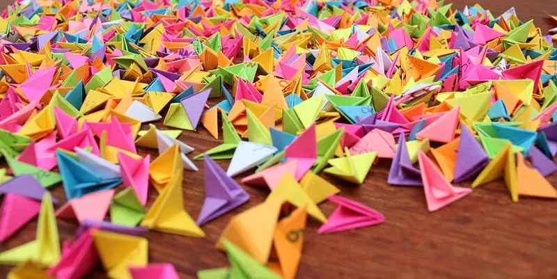 Centenas de post-its coloridos dobrados em pequenos triângulos de origami espalhados sobre uma mesa de madeira