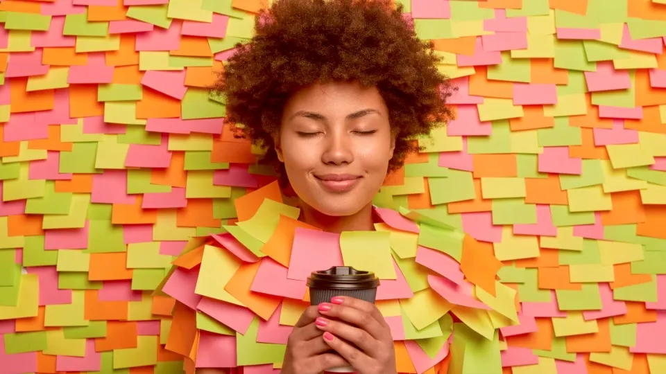 Mulher de cabelo cacheado e olhos fechados, sorrindo, com as mãos em volta de um copo de café, cercada por notas adesivas coloridas
