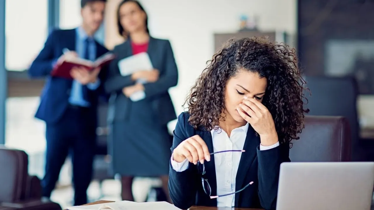Mulher sentada à mesa do escritório, com expressão de cansaço e a mão no rosto, enquanto dois colegas conversam ao fundo olhando para ela
