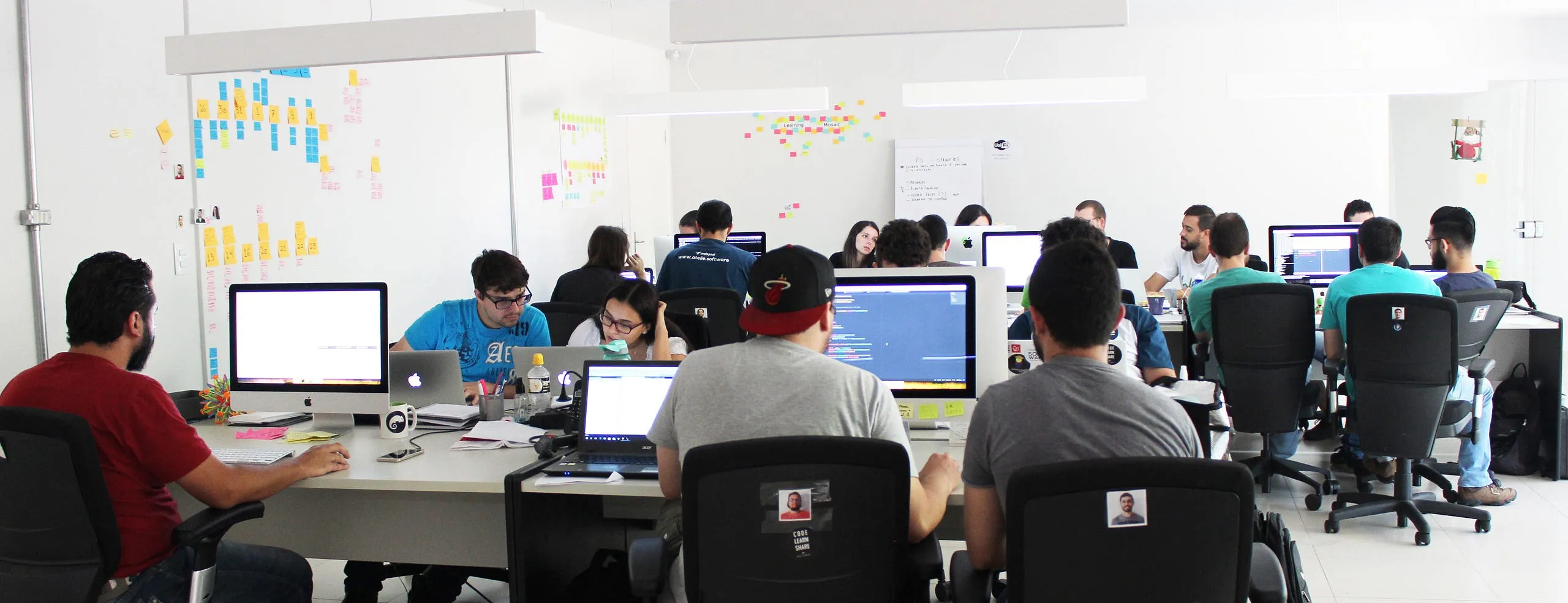 Equipe da Webgoal trabalhando em mesas compartilhadas num escritório aberto, com paredes cobertas de post-its