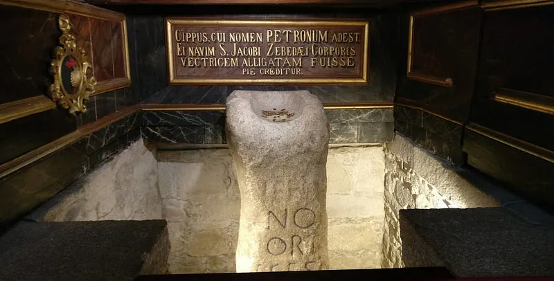 O pedrón, uma coluna de pedra com inscrição romana, sob uma placa em latim na Igreja de Santiago de Padrón